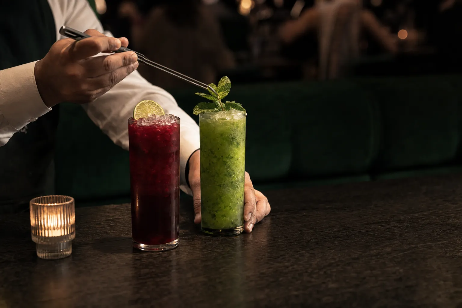 A bartender garnishes a mint lemonade beside a hibiscus drink at a lounge counter.